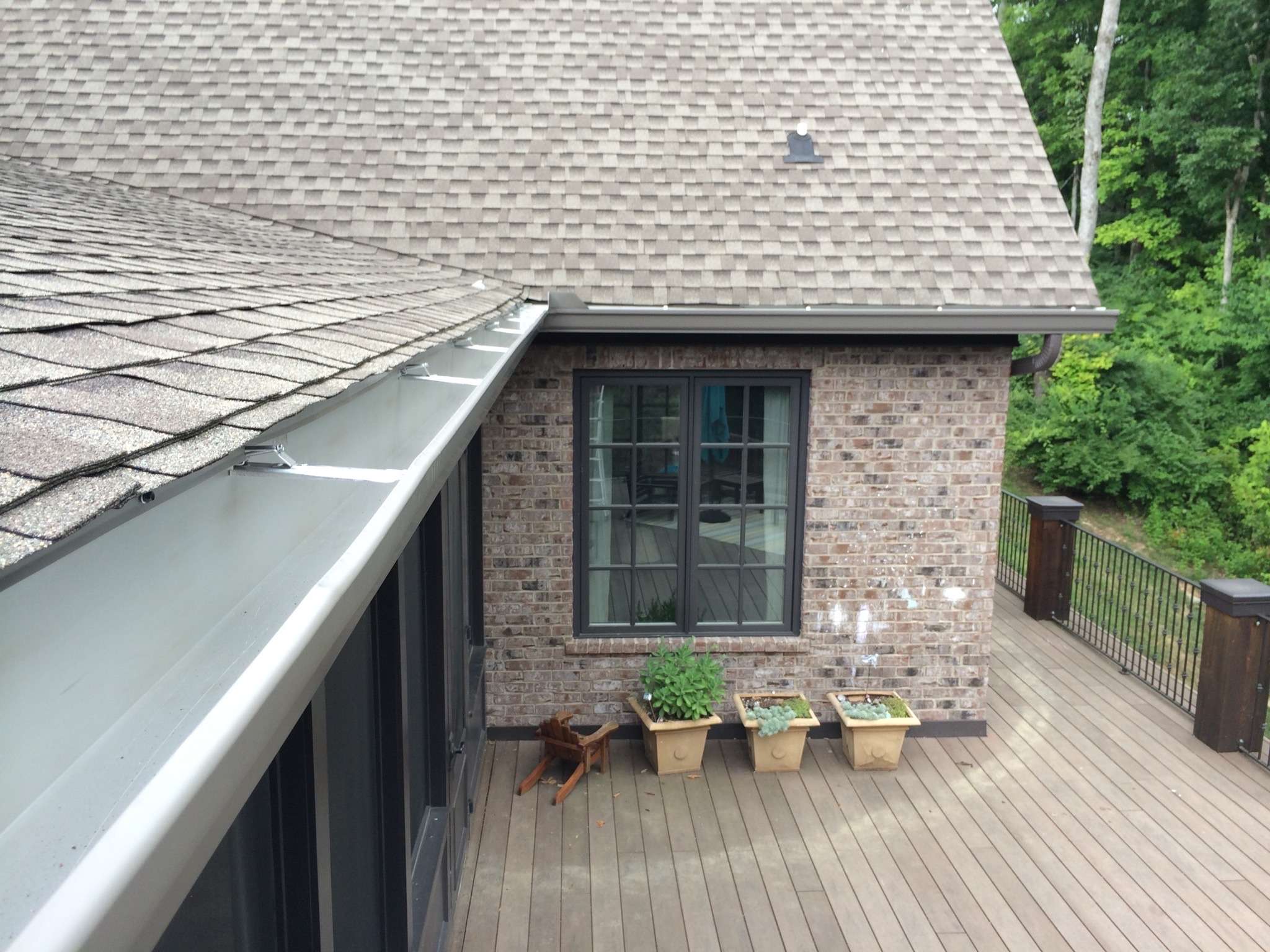 7-inch seamless aluminum gutter in a light bronze color installed on a light brown brick home located above a wooden deck with flower planters below the casement window